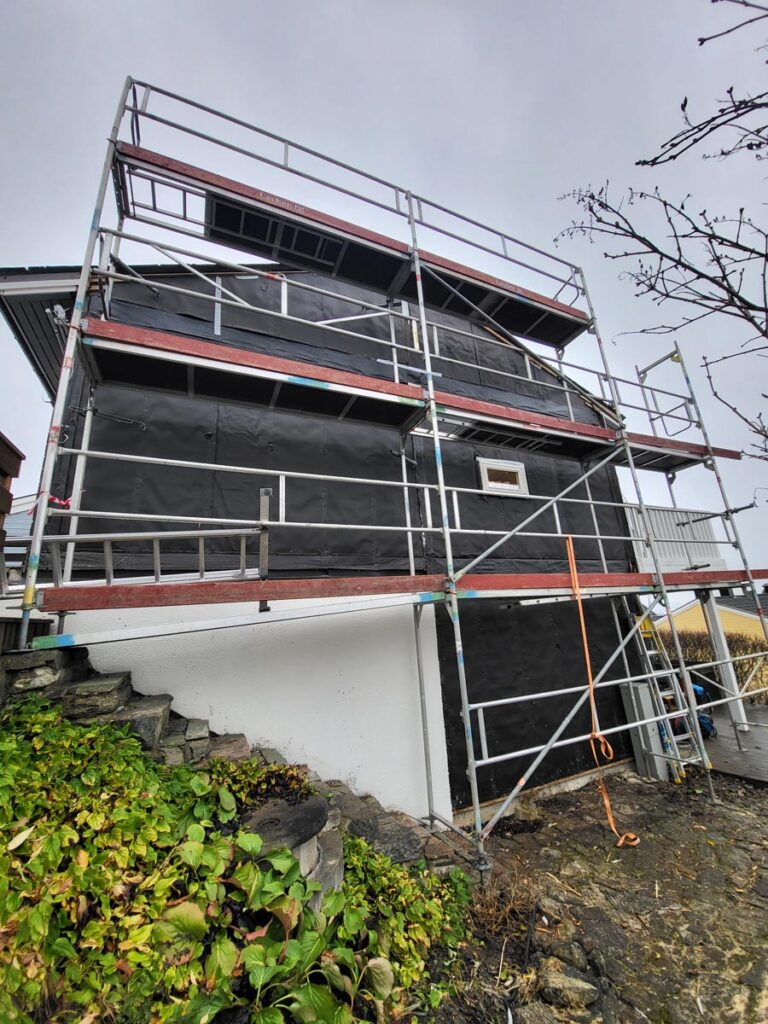 Villa Fjordutsikt, et hus som er under renovering, er innhyllet i svart presenning med hvite nedre vegger. Det er omgitt av stillas og et oransje tau, mens steintrapper og grønne planter gir forgrunnen en sjarmerende stemning.