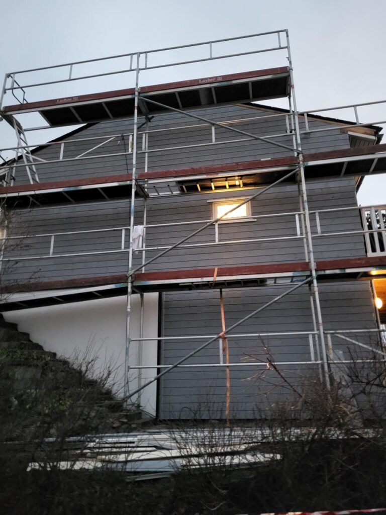 Et grått trehus, Villa Fjordutsikt, er under renovering og omgitt av metallstillas. Ett vindu og en utendørslampe er tent. Det ligger noen planker og byggematerialer på bakken foran huset.