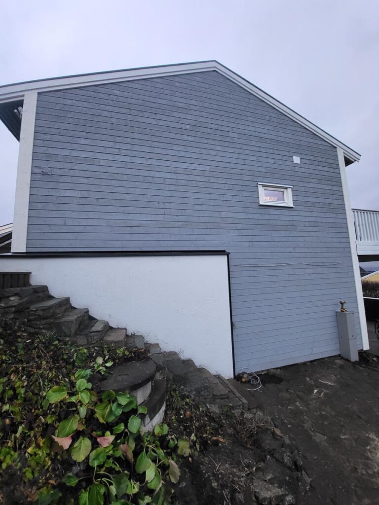 Et blått trehus, Villa Fjordutsikt, med et lite vindu, steintrapper omgitt av grønne planter og en hvit grunnmur. Det ser ut til å være overskyet.