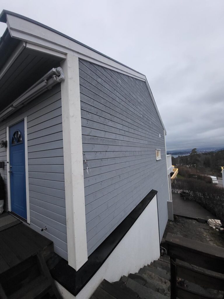 Et sidebilde av Villa Fjordutsikt, et grått trehus med blå inngangsdør, hvite lister og små vinduer. En trapp går langs siden av bygningen. Himlen er overskyet, med trær og hus i bakgrunnen.