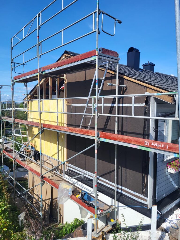 Et hus som er under renovering, muligens Villa Fjordutsikt, er delvis dekket av stillas. Den ene siden er avdekket, slik at man kan se den gule isolasjonen, mens resten er kledd med brune paneler. Himmelen over er klar og blå.