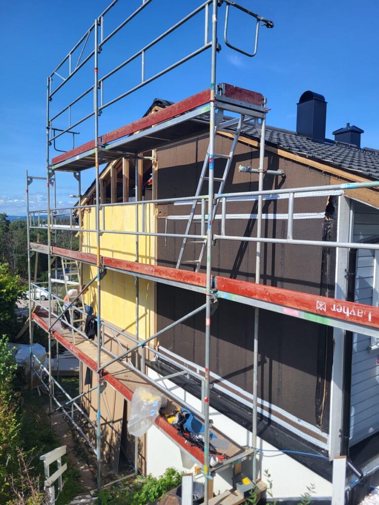 Det står stillas rundt siden av Villa Fjordutsikt, som er under renovering. En del av ytterveggen er blottlagt, slik at isolasjon og byggematerialer er synlige, og det ligger verktøy og utstyr på stillasplattformene.