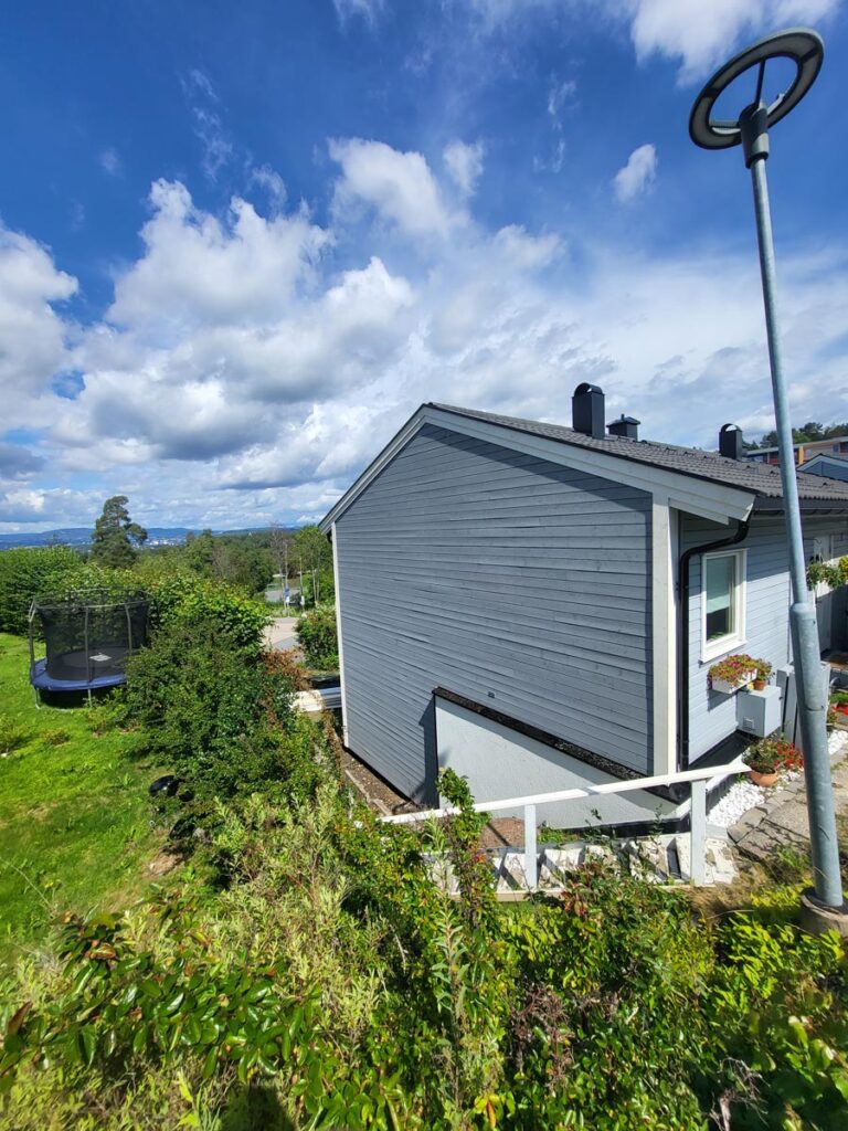 Et lyseblått trehus, Villa Fjordutsikt, med hvite lister, ligger omgitt av grønne busker og gress under en delvis overskyet himmel. Det står en trampoline i bakgården og en gatelampe i forgrunnen.
