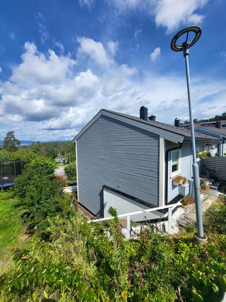Et toetasjes grått hus, Villa Fjordutsikt, med hvite detaljer, ligger omgitt av grøntområder og busker under en delvis skyet blå himmel. En høy gatelampe står i nærheten, og fjellene skimtes i det fjerne.