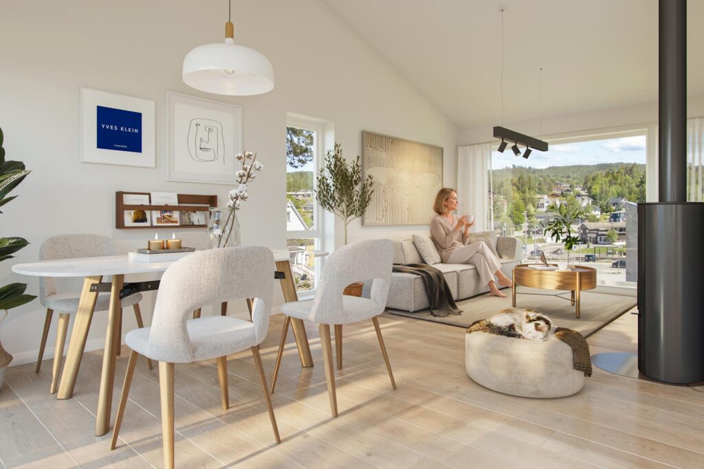 Et lyst og moderne stue- og spisestueområde i Sveitservillaen på Nordstrand, med store vinduer, lyse tregulv, nøytrale møbler, en kvinne som slapper av på sofaen med en kopp i hånden, og en katt som sover på en pute ved siden av en svart ovn.