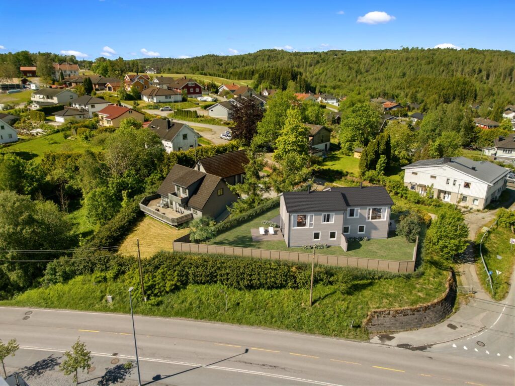 Luftfoto av et forstadsområde med hus omgitt av grøntområder, trær og plener på en solrik dag; hovedveien går forbi Sveitservillaen på Nordstrand, med skogkledde åser i bakgrunnen.