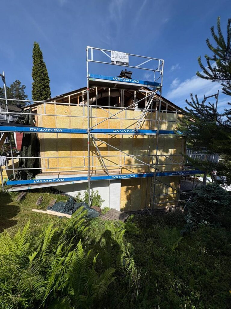Villa Fjordutsikt er under renovering, med stillas langs ytterveggen som er dekket av gul isolasjon. Frodige planter og grøntområder fyller forgrunnen, mens en klar blå himmel strekker seg over det hele.