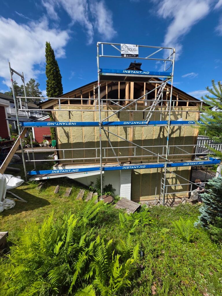 Villa Fjordutsikt er under renovering og omgitt av stillaser. Den øvre fasaden viser synlig isolasjonsmateriale, med grønne planter og et hvitt gjerde i forgrunnen under en delvis overskyet himmel.