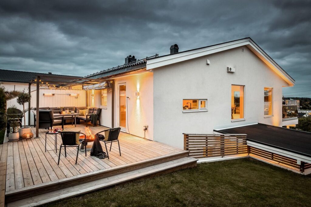 Et moderne hvitt hus med store vinduer og en treterrasse, utstyrt med uteservering, et bord med stoler, lyskjeder og en bålplass, fotografert i skumringen med varm belysning både inne og ute.