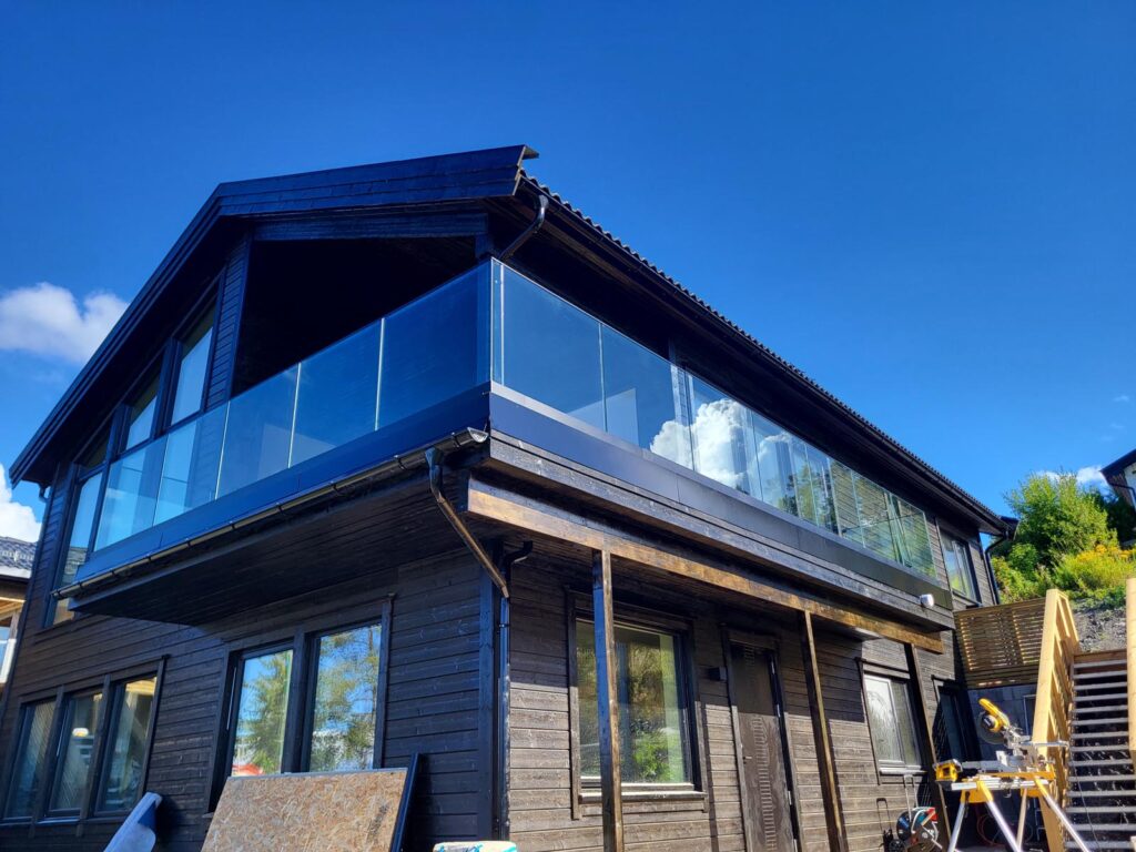 Et toetasjes hus med mørk trebekledning, en stor glassbalkong i øverste etasje og et skråtak under en klarblå himmel. Byggematerialer og trapper er synlige på høyre side.