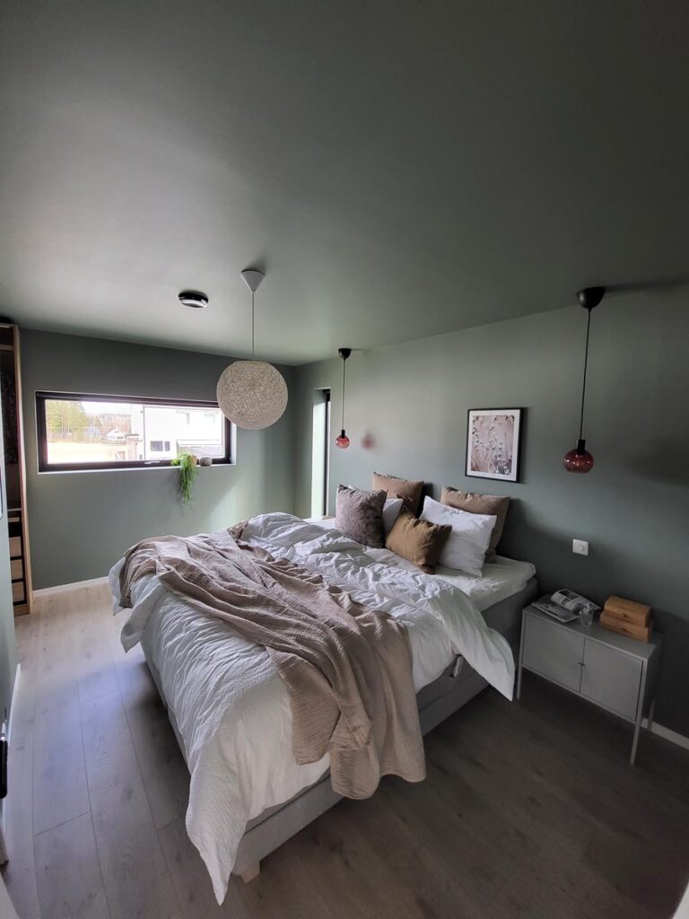 Et moderne soverom med lysegrønne vegger, en dobbeltseng med hvitt og beige sengetøy, to hengende nattbordslamper, et nattbord, et innrammet bilde, et vindu med naturlig lys og en plante i vinduskarmen.