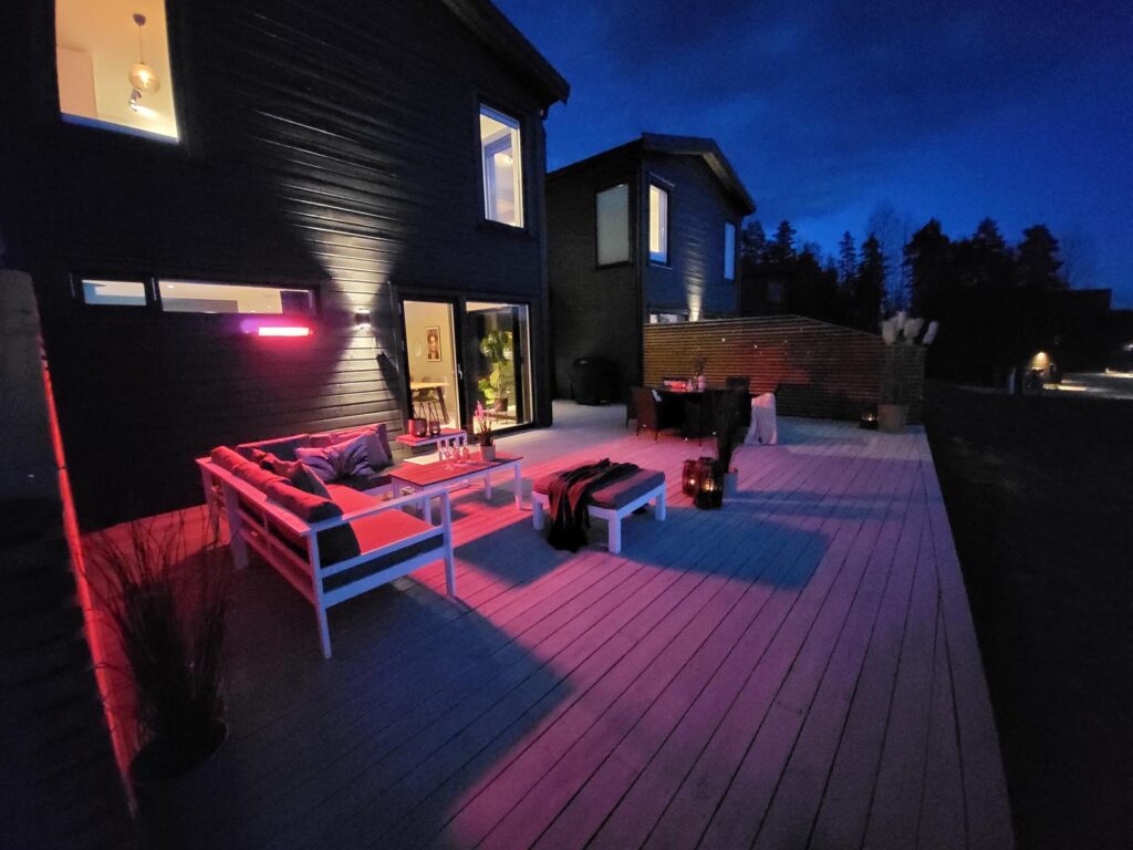 En moderne uteplass om kvelden med loungemøbler, et bord og rosa belysning. Uteplassen ligger i tilknytning til et mørkt hus med store vinduer, og området rundt er svakt opplyst, med trær i bakgrunnen.
