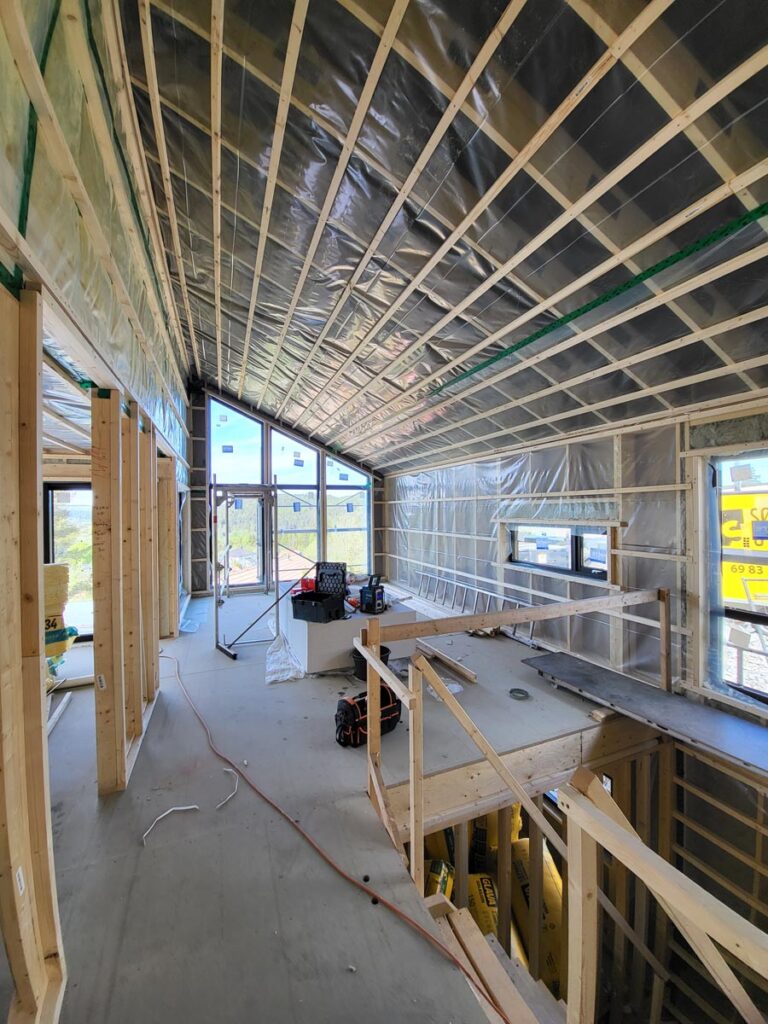 Innvendig utsikt fra et hus under oppføring med synlig trekonstruksjon, delvis isolering, plastfolie, verktøy og arbeidsutstyr spredt utover gulvet. Sollys strømmer inn gjennom store vinduer til venstre.