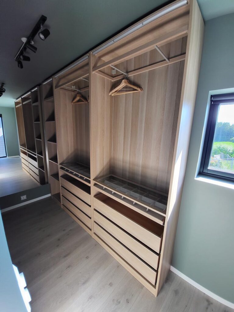 Et moderne walk-in-garderobe med trehyller, skuffer og stativer til å henge klær på. Man kan se noen få kleshengere av tre, og sollyset strømmer inn gjennom et vindu i nærheten. Garderoben er tom, ryddig og velorganisert.