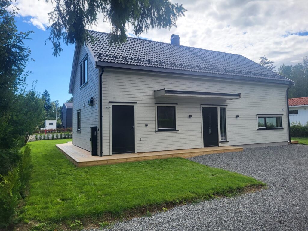 Et moderne toetasjes hus med lysegrå trepanel, svarte takstein og store vinduer. Det har en liten treterrasse, en grusvei og er omgitt av en velstelt plen og trær under en delvis overskyet himmel.
