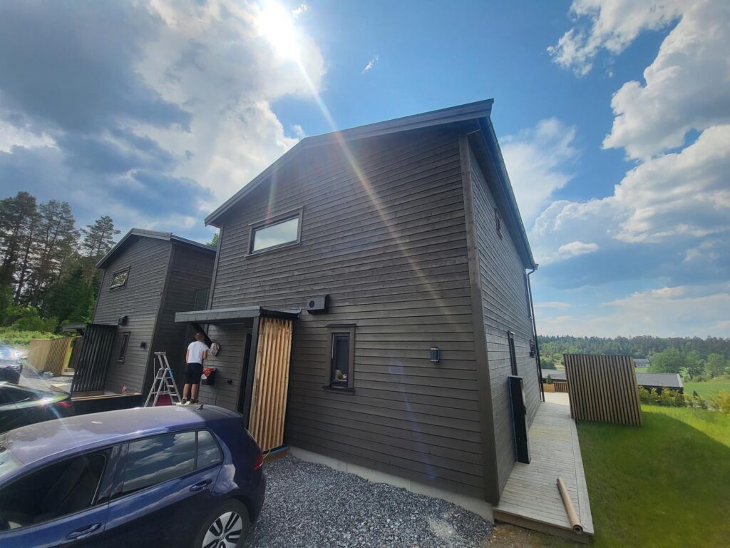 Et moderne hus i mørkt treverk med store vinduer under en lys, delvis overskyet himmel. En person står ved inngangsdøren på en stige, med en parkert bil i nærheten og trær i bakgrunnen.