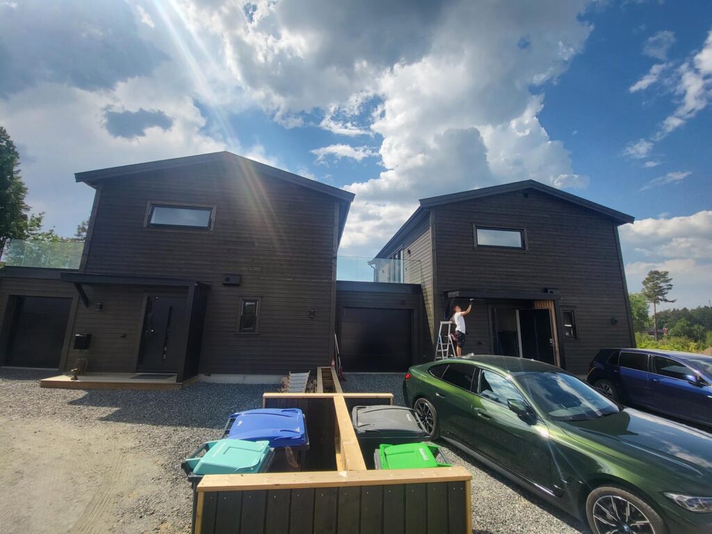 To moderne hus i mørkt tre står side om side under en delvis overskyet himmel. En person på en stige vasker et vindu. Fargerike søppelkasser og parkerte biler, blant annet en grønn bil, står foran et treverkstak. Sollyset skaper reflekser i linsen.