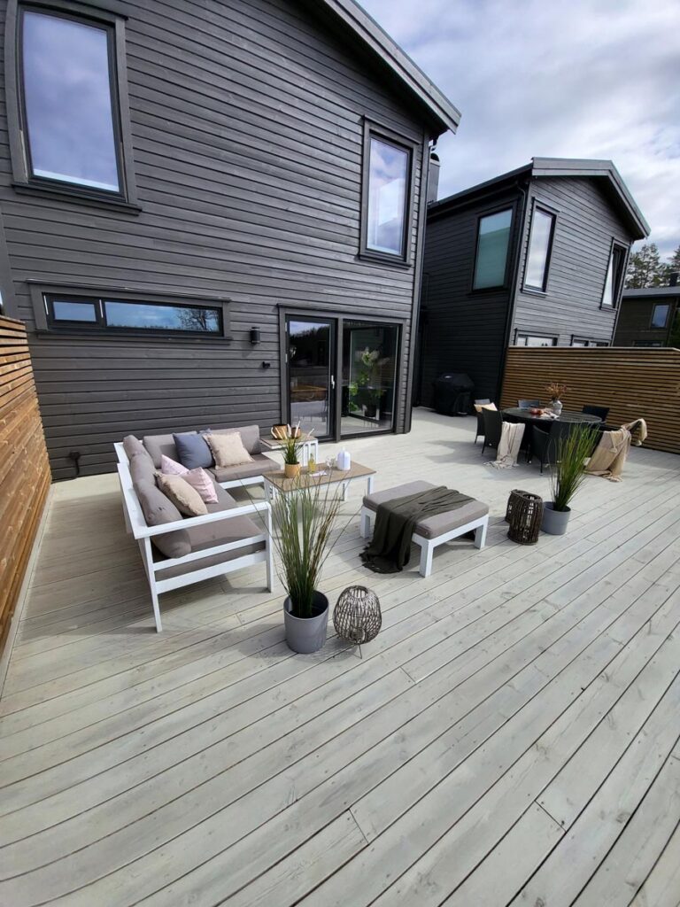En moderne uteplass med lyse treplanker er innredet med en hvit modulsofa, puter, et salongbord, potteplanter og et spiseområde. Huset har mørk fasadekledning, store vinduer og et treverkstak som gir privatliv.