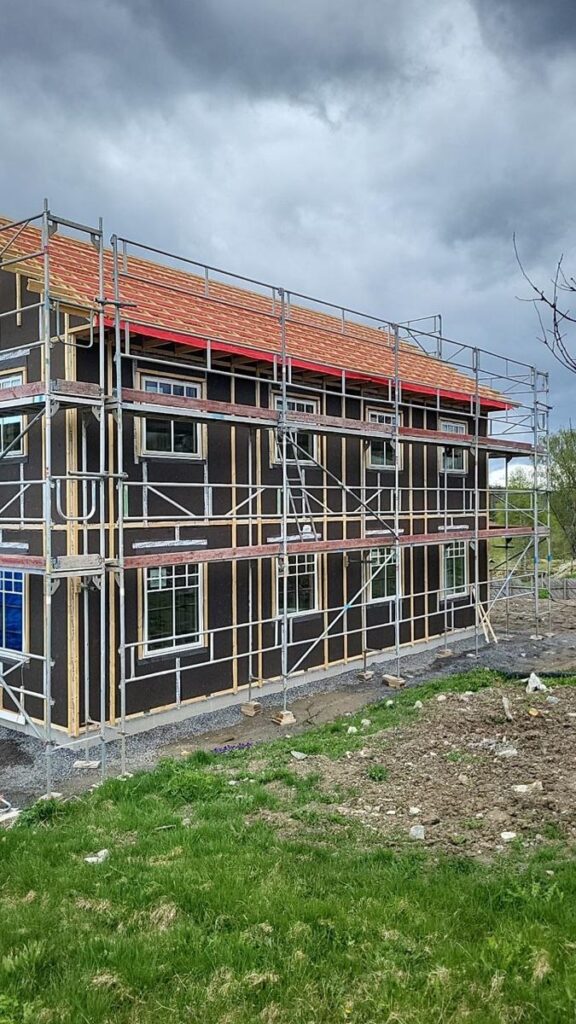 Et toetasjes hus under oppføring, omgitt av metallstillas, med synlig bjelkverk og delvis ferdig tak under en overskyet himmel. Marken rundt huset er bar og gresskledd.