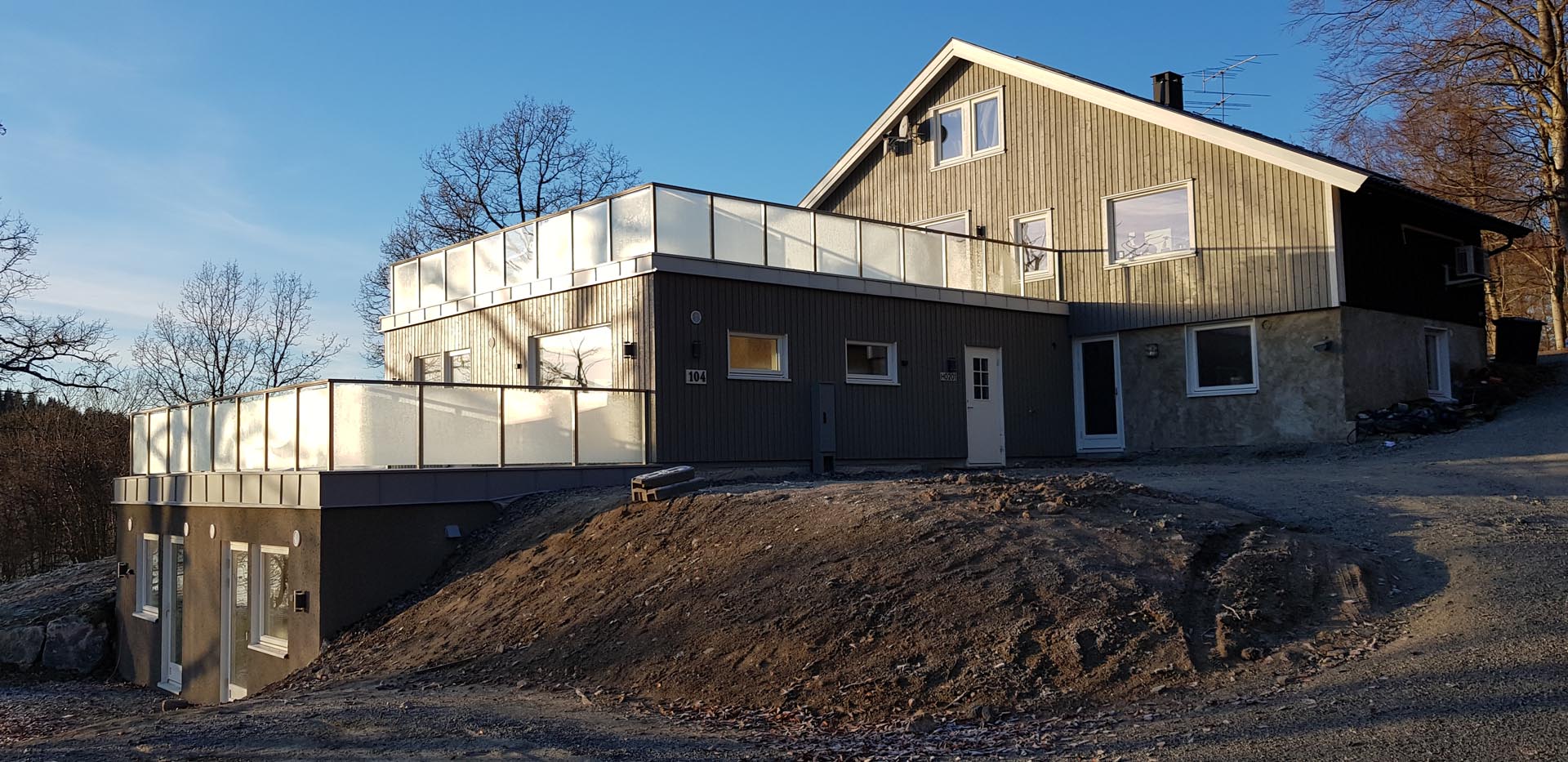 Moderne toetasjes hus med store glassrekkverk på balkongen og trepanel, beliggende på en skrånende, grusbelagt tomt med nakne trær i bakgrunnen under en klar himmel.