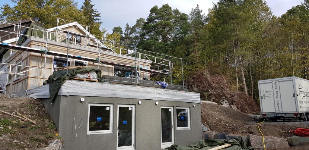 Et hus under oppføring på en skrånende, skogkledd tomt. Den øvre etasjen har stillas og synlige bjelker, mens den nedre etasjen har en ferdig utvendig fasade med tre dører. Byggematerialer og en hvit anleggsvogn står i nærheten.