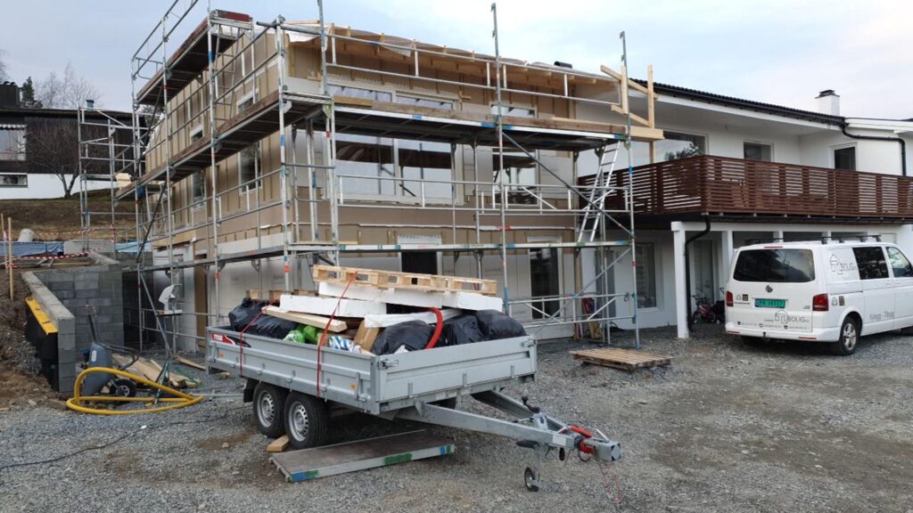 Et hus under oppføring med stillas rundt seg, byggematerialer lastet på en tilhenger foran, og en hvit varebil parkert i nærheten på en grusvei.