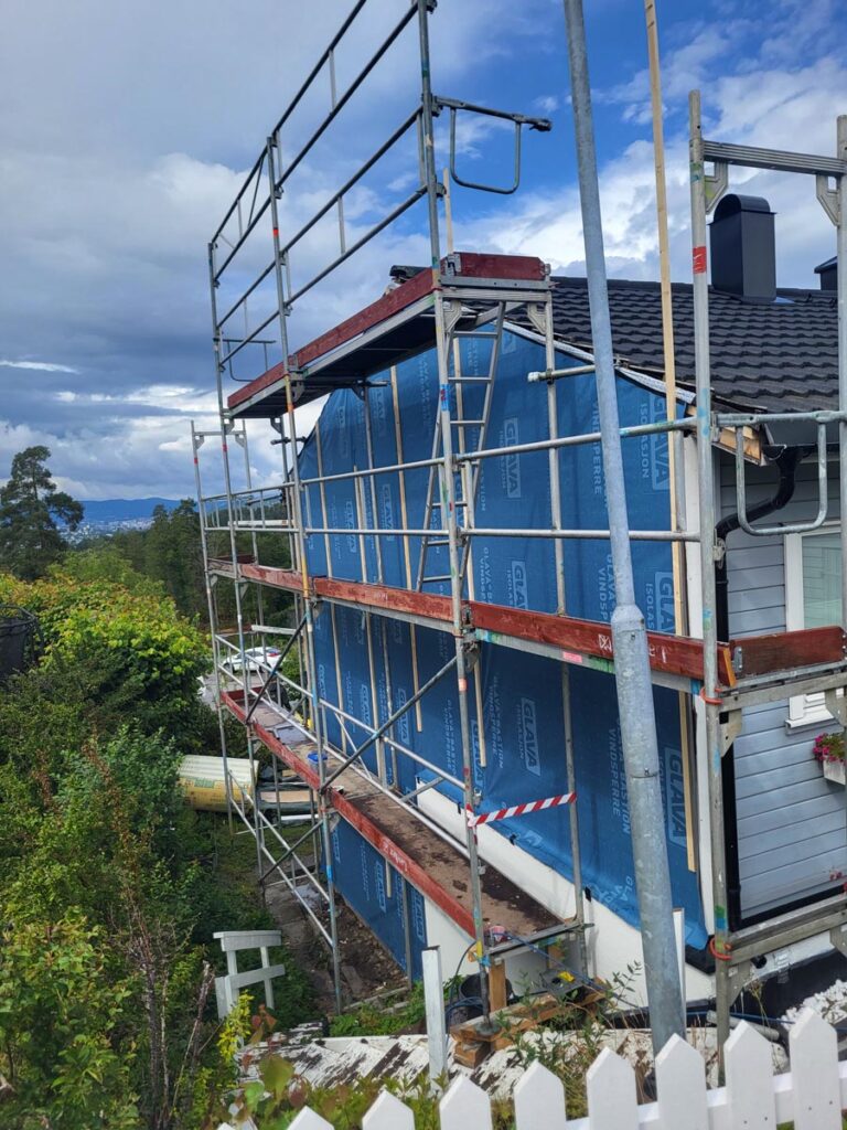 Et hus under renovering med stillas langs siden og blå, værbestandige presenninger som dekker fasaden. Området er omgitt av trær og busker, og i forgrunnen ser man et hvitt stakittgjerde.